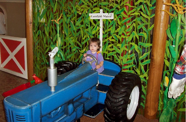 Small child sitting on a child-sized tractor with a cornfield mural backdrop.