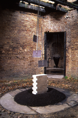 One Day Four Hours: Multi-room, building wide installation in Potsdam Germany by Edward Dormer, featuring 3 suspended shovels over white pristine bricks emerging from a ring of coal dust in a deteriorated brick structure, examining institutional memory. 