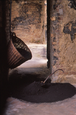 Wicker basket hanging on a wall in a doorway filled with bituminous coal dust, with a rusted shovel whose wooden handle has been replaced by burnt oxidized copper wires curved downwards, the blade dug into a pile of coal dust beneath the overflowing basket.