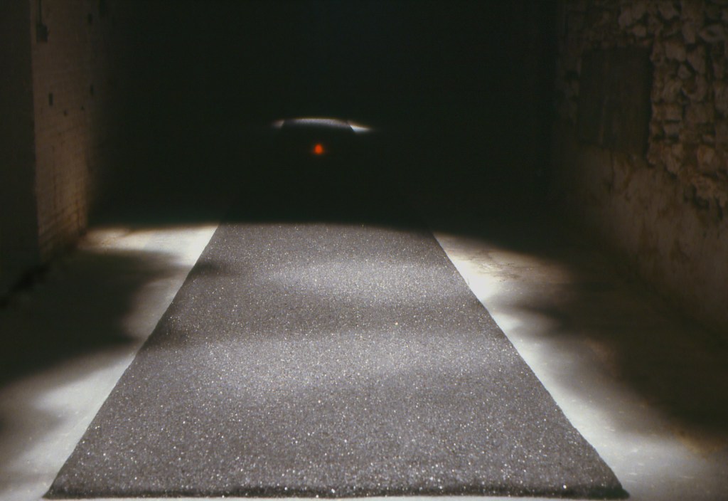 "View through installation space showing laser-proton elements on darkened gallery floor."
