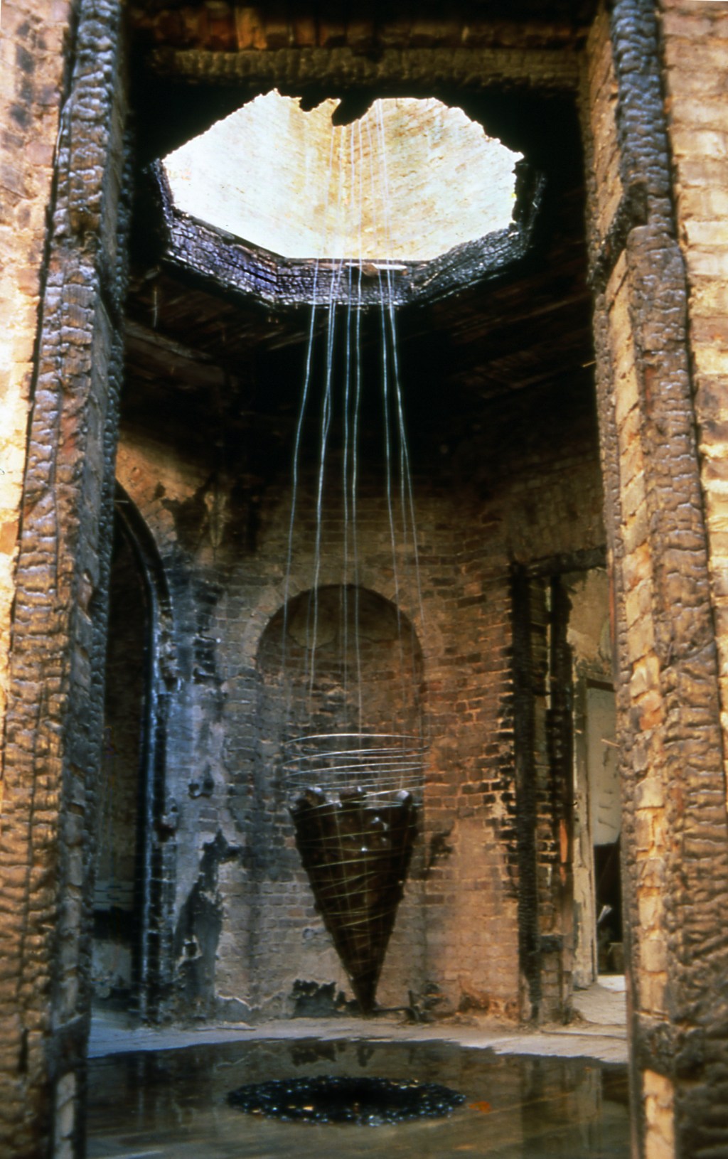 One Day Four Hours: Pendulum installation by Edward Dormer at Schiffbaubauergasse Potsdam, showing a cone-shaped wire structure suspended over bituminous coal in a former East German military base, exploring themes of destruction and renewal.